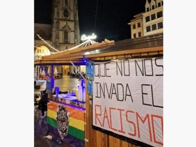 Caseta de les Fiestes Populares d’Uviéu con una bandera arcoíris y una pancarta contra’l racismu.