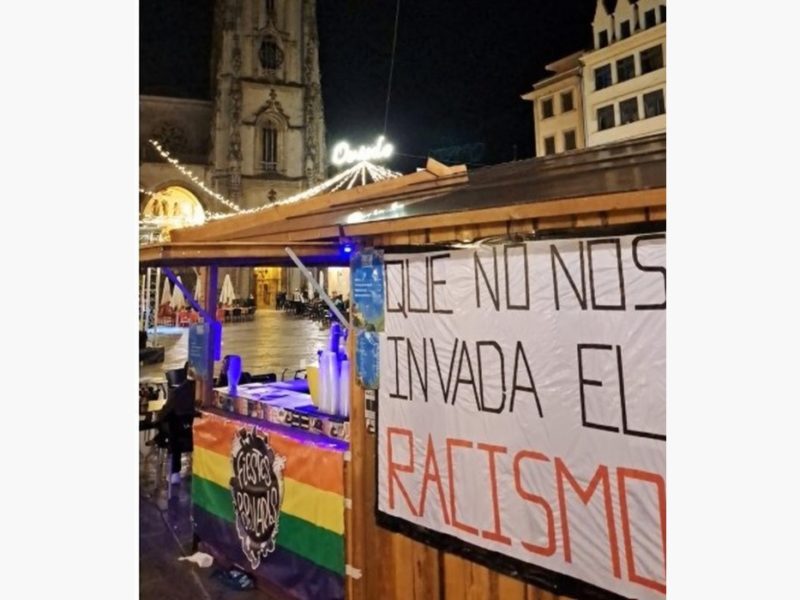 Caseta de les Fiestes Populares d’Uviéu con una bandera arcoíris y una pancarta contra’l racismu.
