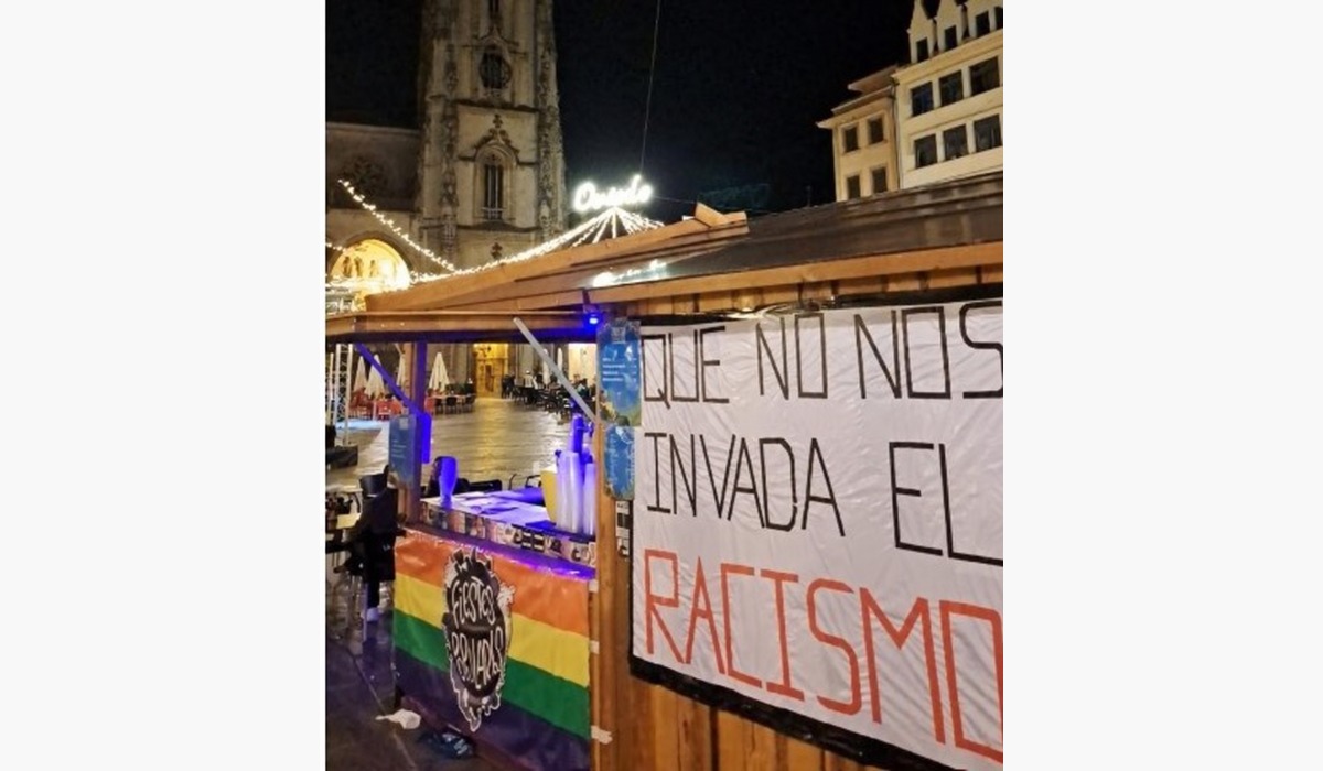 Caseta de les Fiestes Populares d’Uviéu con una bandera arcoíris y una pancarta contra’l racismu.