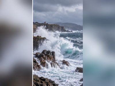 Foles rompiendo contra un llitoral pedregosu; imaxe xenerada con IA.