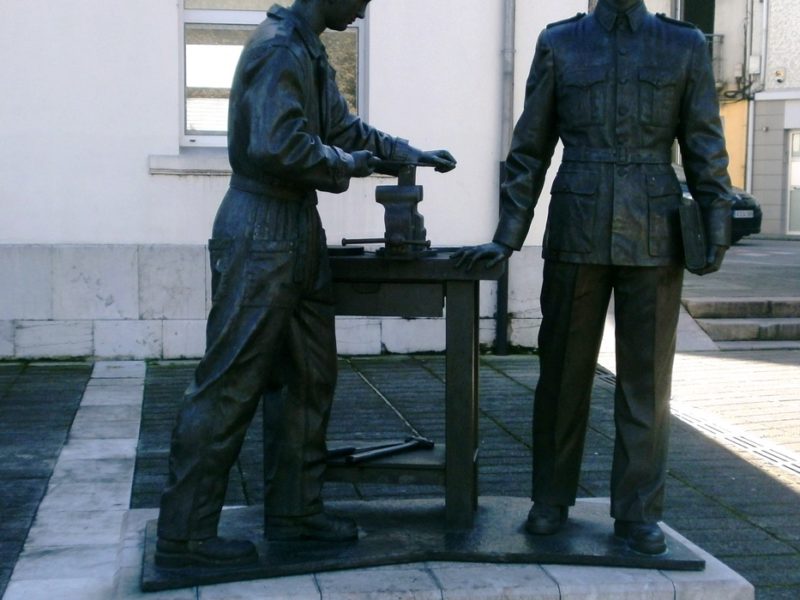 Escultura de bronce con dos figures de trabayadores xunto a un bancu de taller en Trubia.