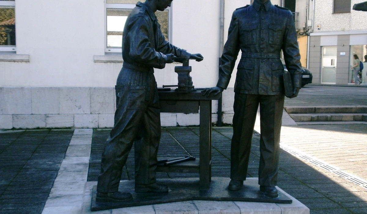 Escultura de bronce con dos figures de trabayadores xunto a un bancu de taller en Trubia.